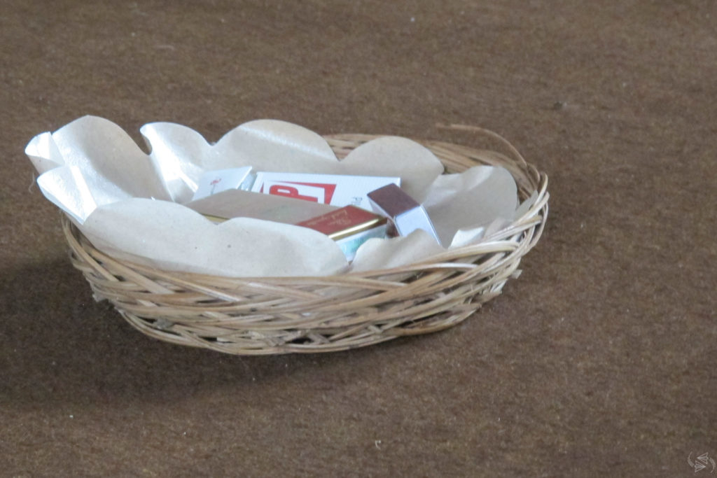 A thatched shallow container on the carpeted floor, containing packs of various brands of cigarettes.