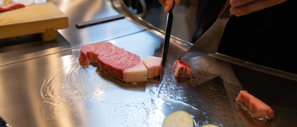 A kobe beef steak is being cut into precise pieces on the hot plate with a knife and metal spatula.