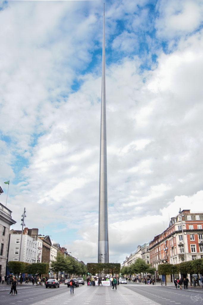 The Spire in Dublin. The shiny silver metal monument is larger at the base and gets gradually smaller as it goes up, like a pointy stick. The Spire starts at street level and is shown rising into the sky with a backdrop of clouds. 