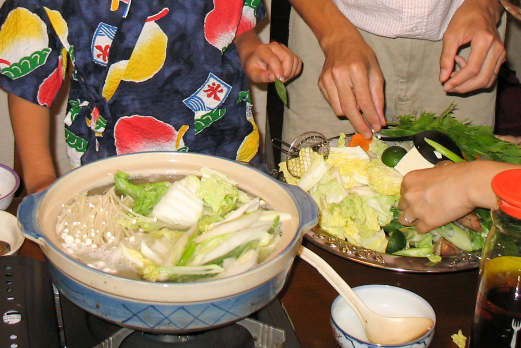 A Japanese family gathers around a table preparing fugu nabe. There is a large nabe pot filled with broth, fugu and vegetables, with a large plate next to it of more vegetables and tofu to add later.