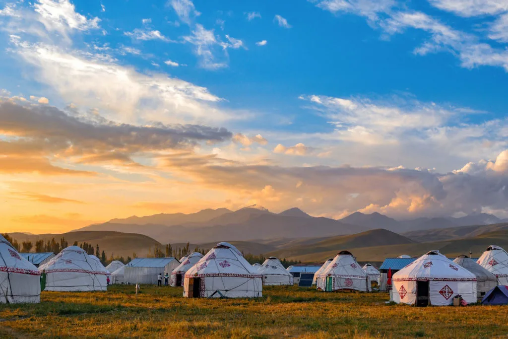 Photo by Yang Shuo (via Unsplash). Numerous white gers with red patterned details on their canvas exteriors stand quite close to one another. In the background, there is a mountain range and the camp is lit up in a golden hour glow.