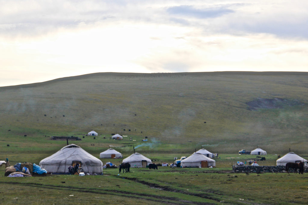 Photo by Bolatbek Gabiden (Via Unsplash). A number of white gers are set up on the plain at the bottom of a hill. Smoke can be seen emanating from the chimneys and several horses are grazing.