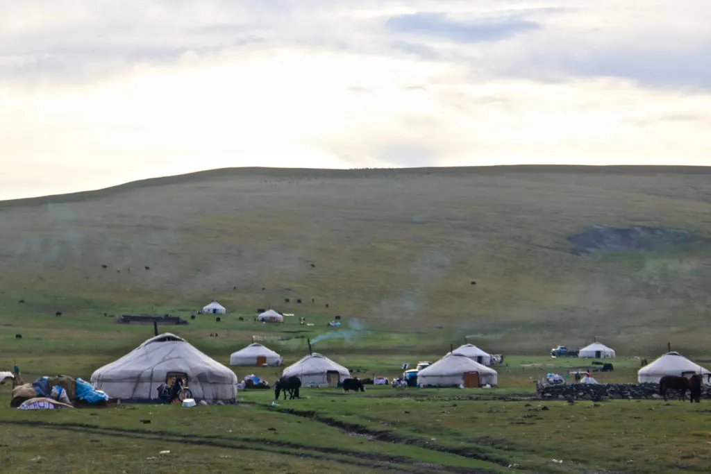 Photo by Bolatbek Gabiden (Via Unsplash). A number of white gers are set up on the plain at the bottom of a hill. Smoke can be seen emanating from the chimneys and several horses are grazing.