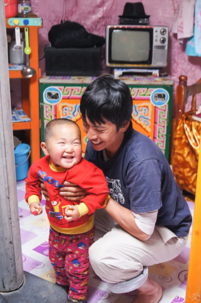 The author's husband laughs with a small boy, who is excited to play games with the guests in his family ger. In the background, shelves with kitchen utensils, a cupboard with a TV on top, and a bed can be seen.