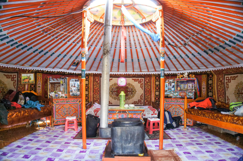 The interior of a ger. The roof and supporting poles are painted in orange and blue. There's a hearth in the center and beyond that is a dining table with stools. The entire middle section has a purple and white plastic matting underfoot. The walls are lined with impressively designed rugs as well as furniture and beds.