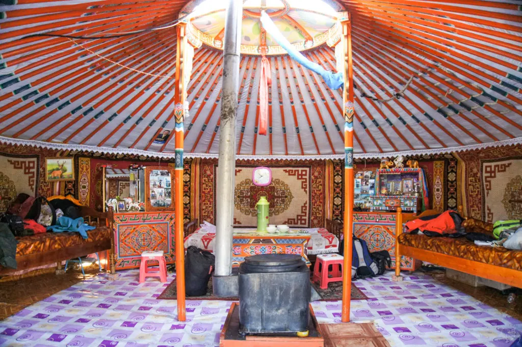 The interior of a ger. The roof and supporting poles are painted in orange and blue. There's a hearth in the center and beyond that is a dining table with stools. The entire middle section has a purple and white plastic matting underfoot. The walls are lined with impressively designed rugs as well as furniture and beds.