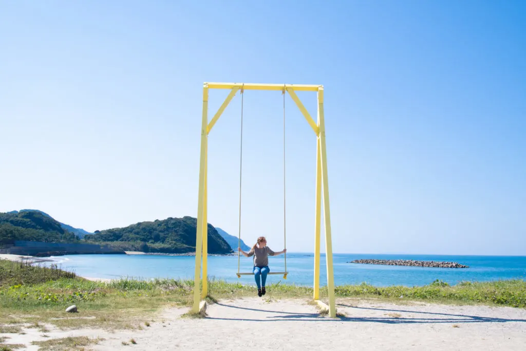 Woman sits on giant yellow swing with the Itoshima coast in the background.