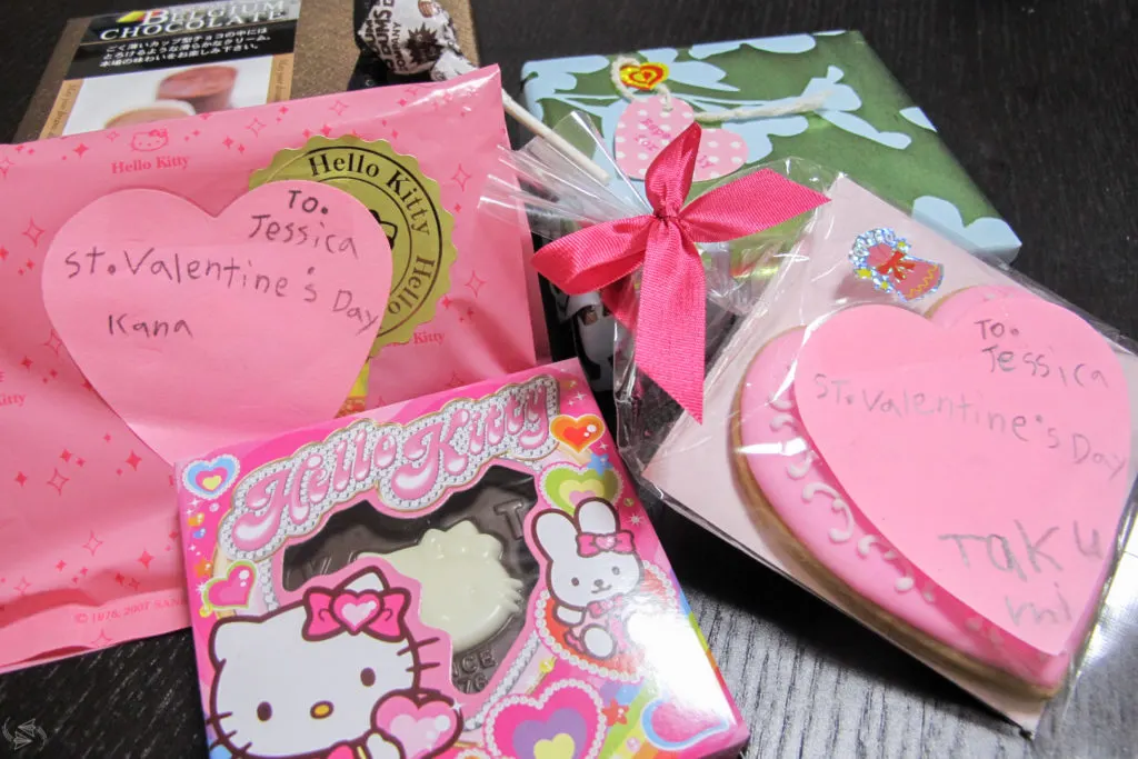 Several examples of giri choco for Japanese Valentine's Day laid out on a table, including a heart-shaped cookie in a bag with a pink bow, a Hello Kitty chocolate heart, small boxes of chocolates and a lollipop.
