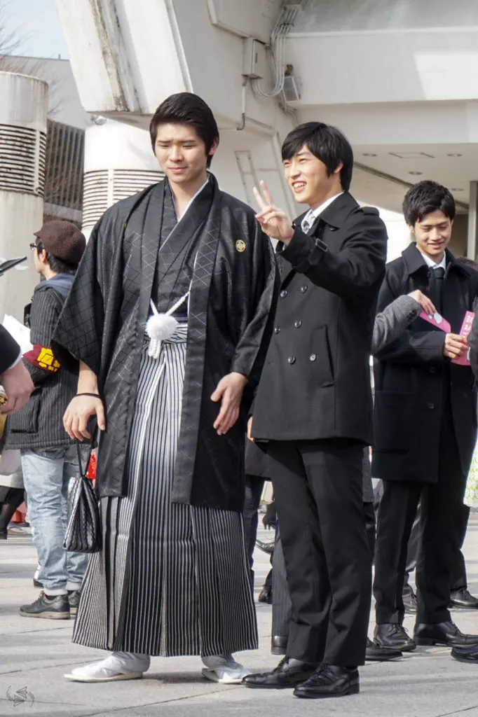 A man wearing traditional hakama poses for a photo next to a man wearing a Western style suit. The man in the suit is giving the peace sign.