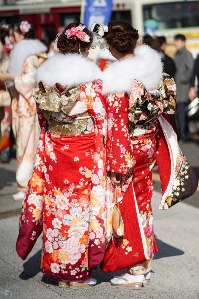 Two women with their backs to the camera, one holding up a smartphone to take a selfie, are all dressed up in their red kimonos with white fur stoles for Japan's Coming of Age Day.