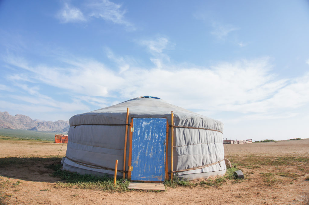 A Mongolian ger in the Semi Gobi Desert with a greyish white canvas and blue door with orange frame.