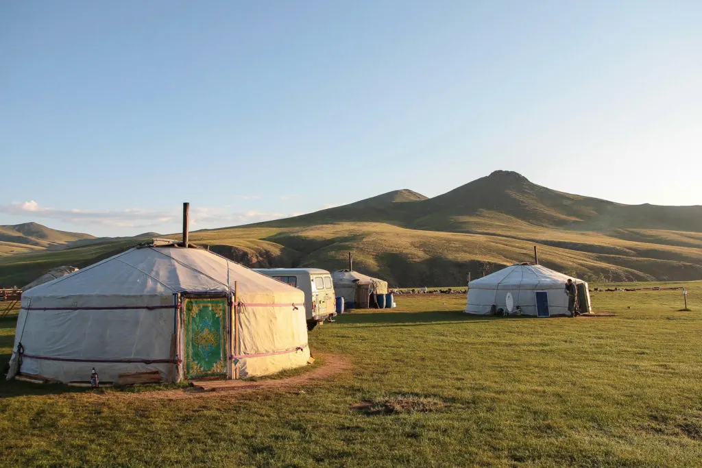 Photo by Vince Gx (via Unsplash). Several Mongolian gers are set up on a grassy plain, the closest with a blue-green door and orange detailing. A white van is parked in the center of the picture.