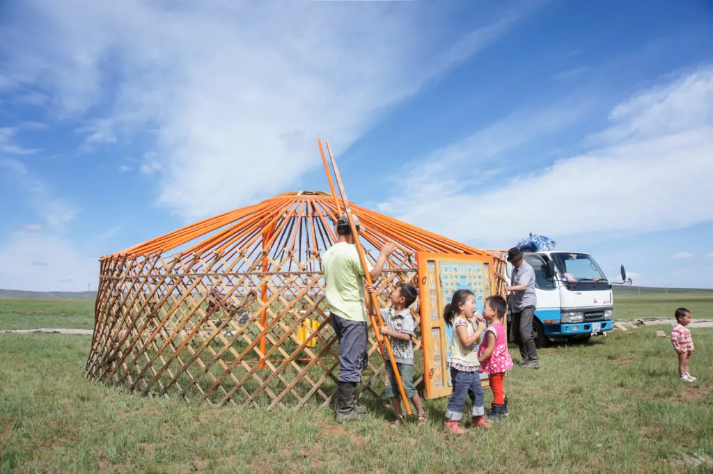 A couple of men and a child put up the last poles and make final adjustments to a ger before getting ready to cover it with felt. Several other children stand around watching, one eating a snack. A pickup truck can be seen to the right of picture with the rest of the ger materials in the back.