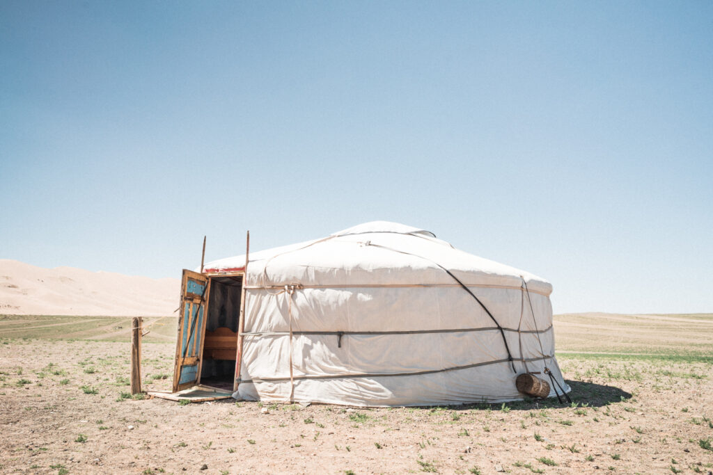 A photo by Patrick Schneider (via Unsplash). A single white ger stands on an arid swathe of land with the door open. The interior door is blue with an orange frame.