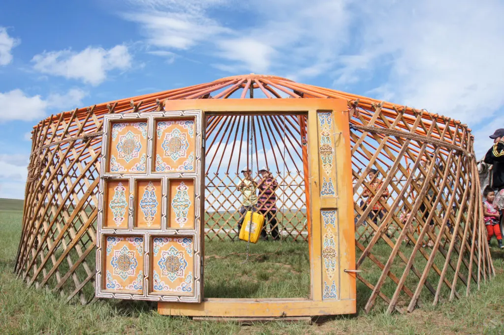 The wooden structure of the ger (poles, lattice and door) have been erected on a grassy plain and await to be covered. The door is wide open revealing a predominanly orange colored design with some intricate details in blue, green and earthy tones.