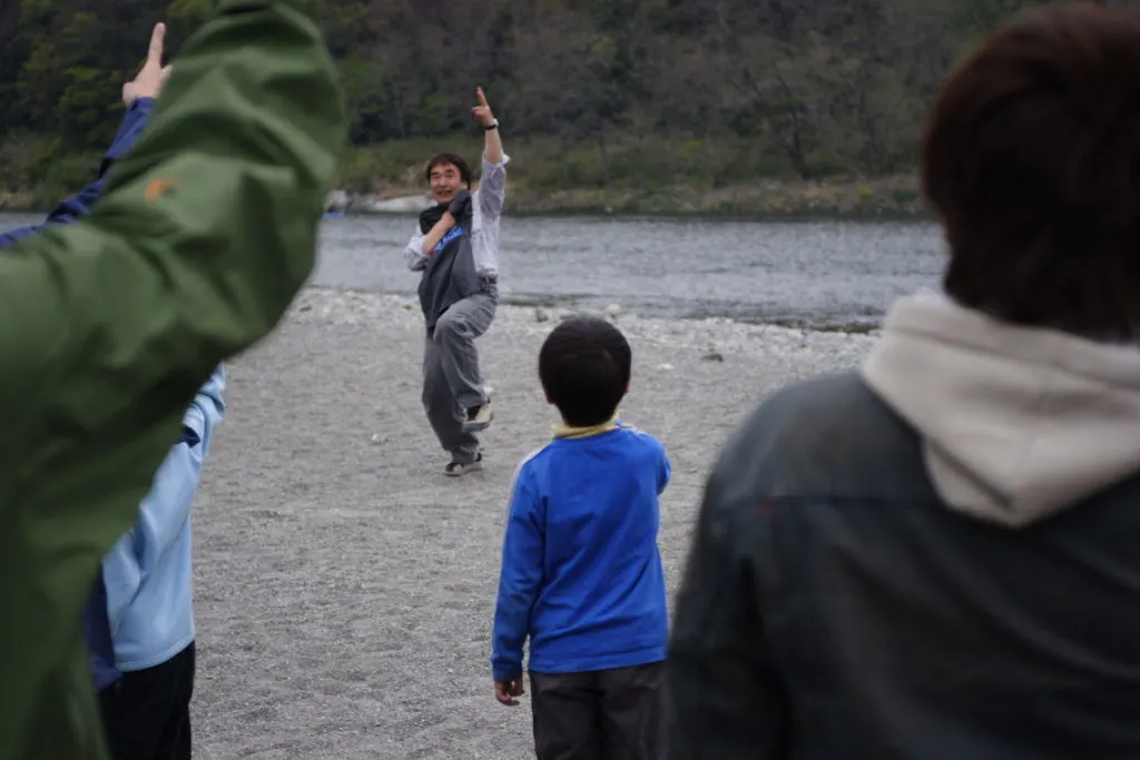 A group play multiplayer janken outdoors by a river. The central player can be seen energetically holding up scissors with his left hand. We can see the backs of four players in the group, holding up their chosen gestures.