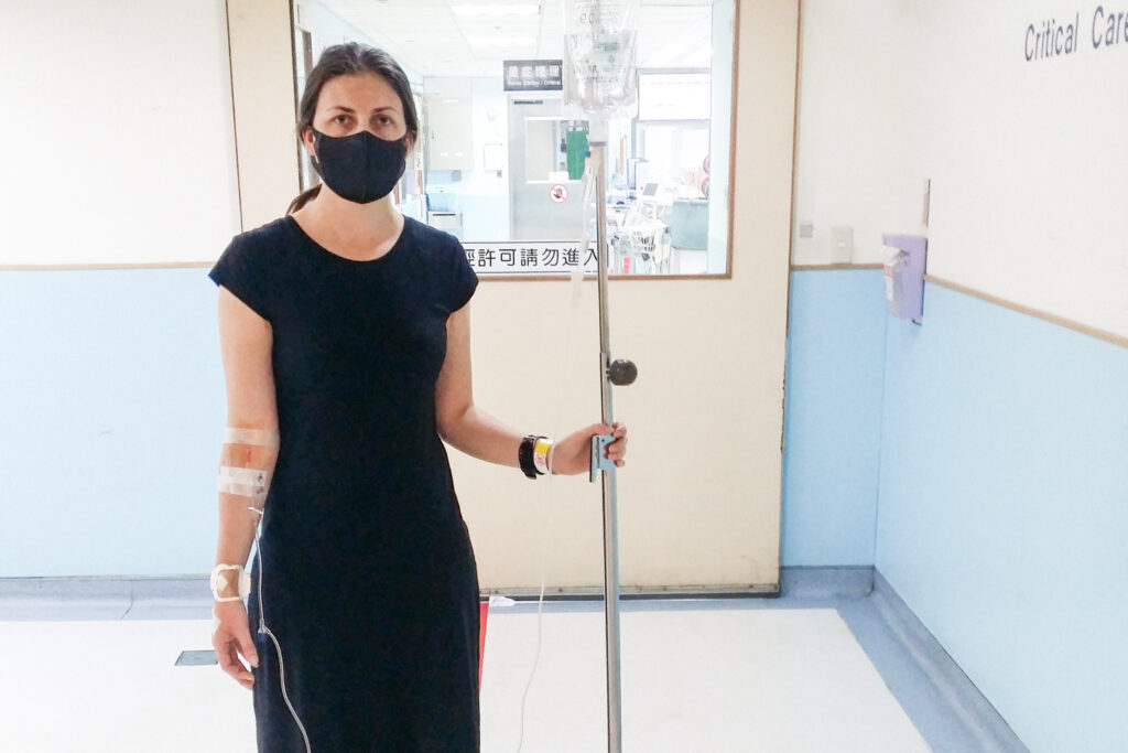 The author stands in a Taiwanese hospital wearing a black face mask with IV drip and stand, and multiple tapes and bandages from blood test attempts.