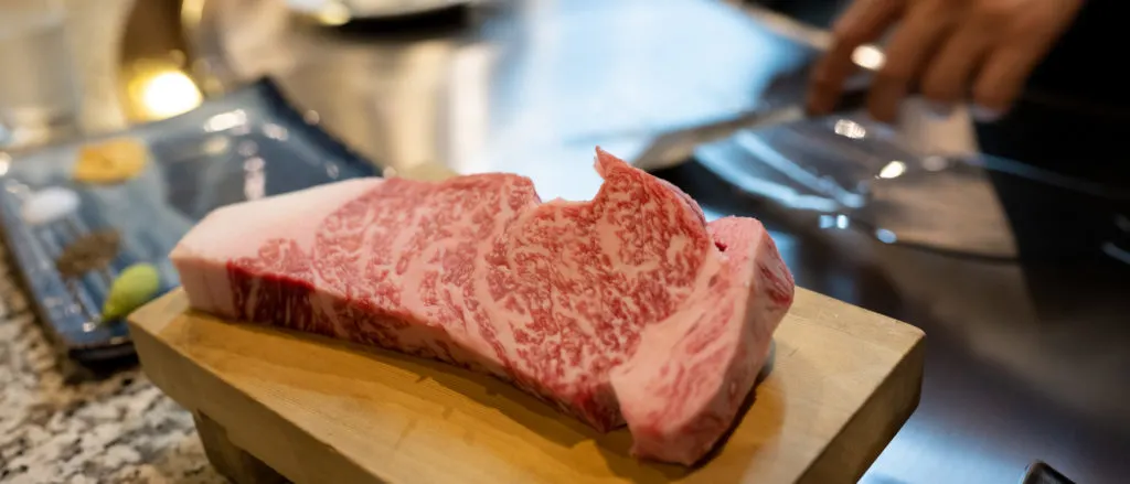 A raw kobe beef steak sits on a wooden chopping board in the foreground while the hands of a chef can be seen preparing the hot plate for cooking in the background.