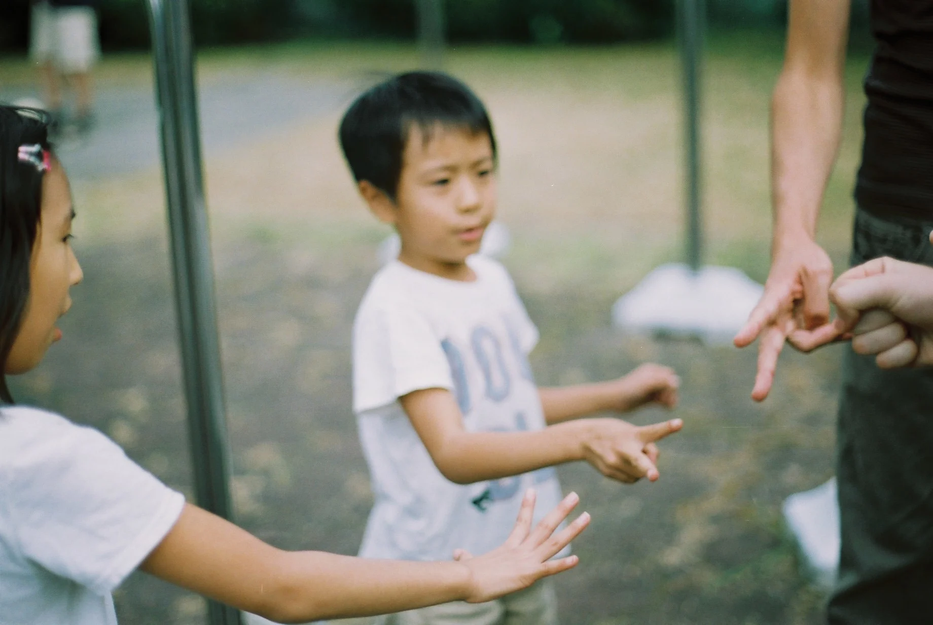 Four people, two children and two adults, play janken against one another outdoors. The girl chose paper, and the boy scissors, while the two adults (faces not shown) chose scissors and rock, resulting in a draw.