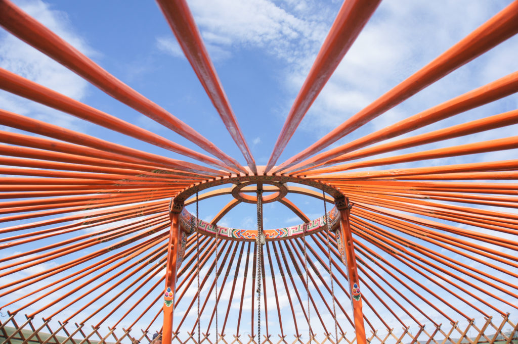 Looking up at the roof of a Mongolian ger before it has been covered with felt. The wooden poles are orange and the sky in the background blue with a few clouds.