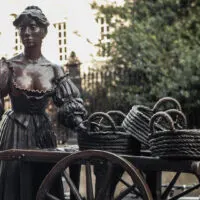 The Molly Statue, Dublin. Molly Malone stands in a low-cut dress with puffy sleeves and long skirt, pushing a wooden trolley with three woven baskets sitting on top.