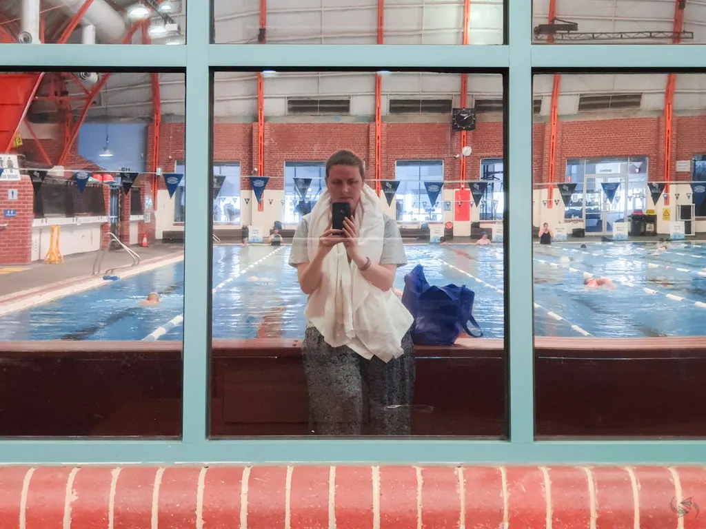 Author takes a post-swim photo of herself reflected in the windows at the indoor swimming pool. Behind her other patrons are doing laps in the pool.