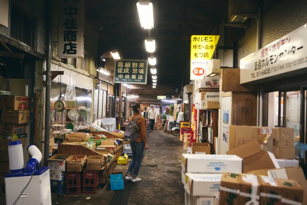 Woman stands in narrow Tanga Market with stores either side and stacked cardboard boxes of produce. 