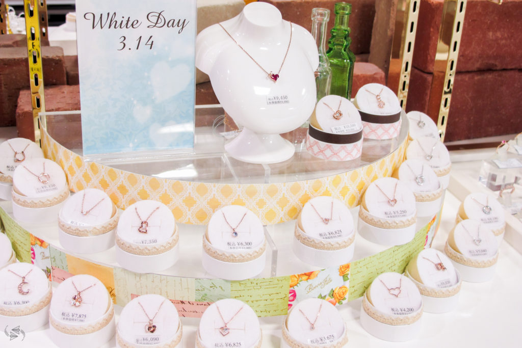 A tiered jewelry display in a department store, showcasing various necklaces, next to a sign that says 'White Day 3.14'.