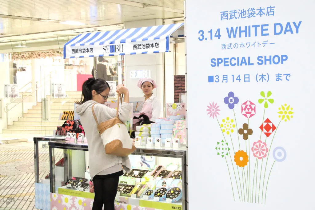 A woman reaches into her purse to get her wallet at a special Seibu department store pop-up White Day shop selling an array of boxed chocolates for the occasion. A big sign to the right of the store says '3.14 White Day Special Shop, until March 14th' with an image of a bunch of flowers underneath.