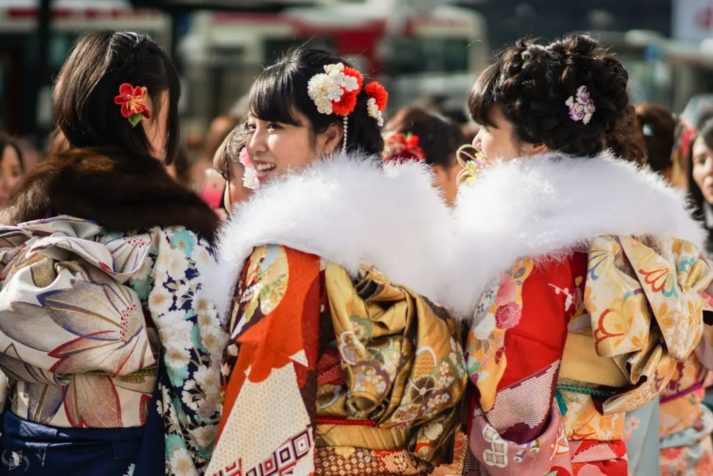 Three Japanese women wearing formal furisode kimono for Seijin no Hi stand together, two facing away, while the one in the middle looks back towards the camera. The one in the middle and the one on the right wear white fur stoles over their striking red, orange and gold kimonos.