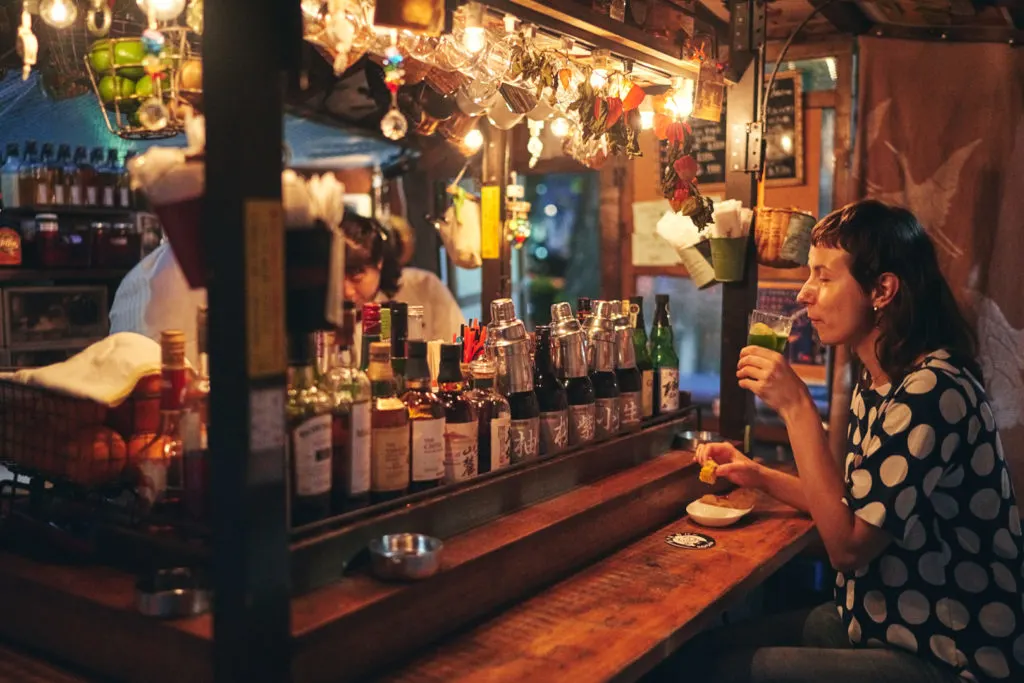 A woman drinks at a fancy bar-type yatai where the counter is lined with various bottles of alcohol.